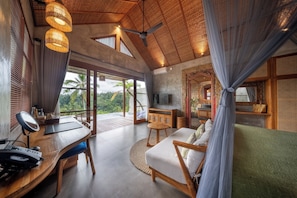 A spacious room with a wooden ceiling, a large window offering a view of palm trees, a sofa, a desk with a computer, and a television.