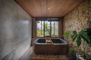 A modern bathroom with a large bathtub, a window with a view of greenery, and a stone wall. - ULU Valley Retreat  (Sebatu)
