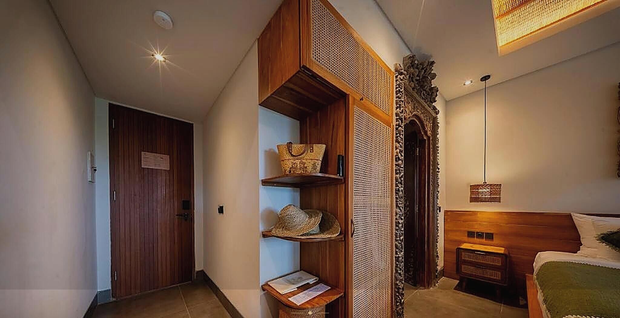 A room with a wooden door, a shelf with hats and a bag, a bed with a headboard, and a wall-mounted light fixture.