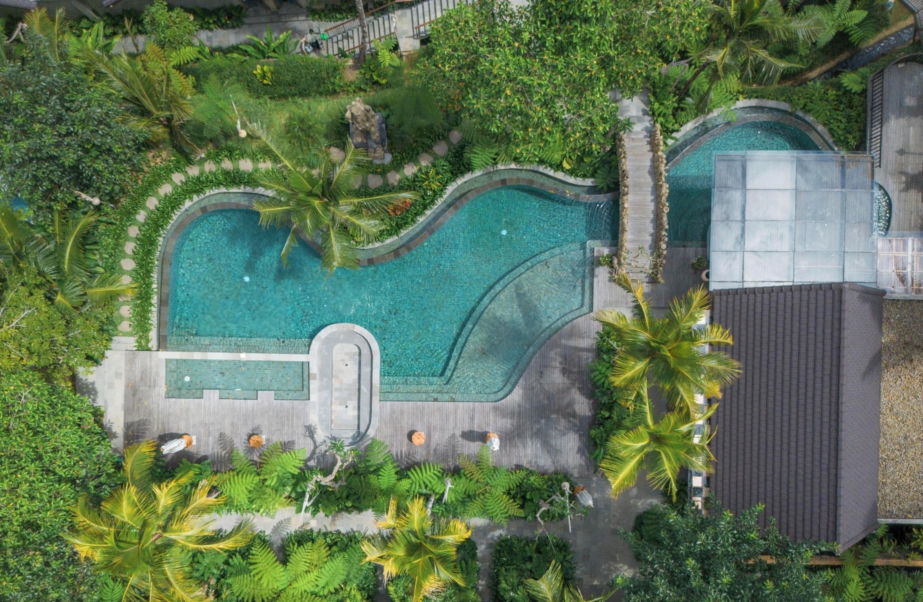 An aerial view of a resort with a large swimming pool surrounded by lush greenery and palm trees.