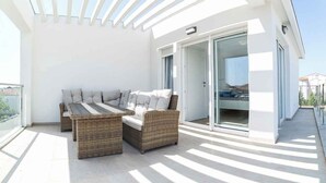 Zona d'estar a l'aire lliure il·luminada per sol, amb moble de vugada, una porta de vidre que dona a un balcó i una façana d'edifici moderna.