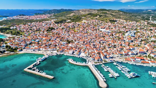 Vista aèria d'una ciutat costanera amb un port esportiu, aigües turqueses cristal·lines i edificis amb teulades vermelles.