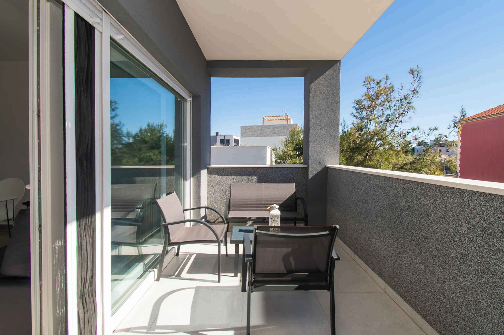 Un balcone con una porta vetrata, un tavolo, due sedie e vista su edifici e alberi.