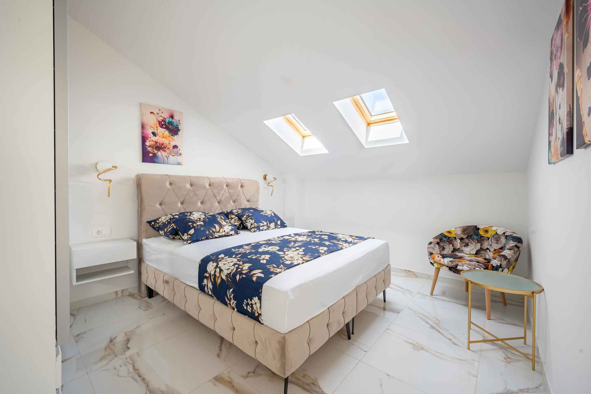 A bedroom with a bed, a chair, a small table, a wall-mounted light, and a ceiling skylight.