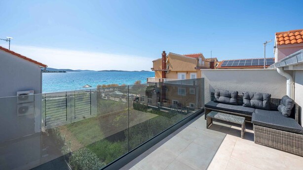 Een balkon met glazen balustrade en uitzicht op de kust, een rieten zithoek met kussens, en een klein tafeltje.