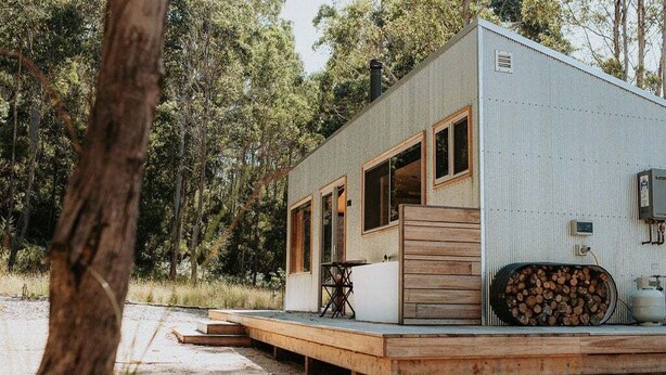 A small cabin with a deck, large windows, and a stack of firewood.