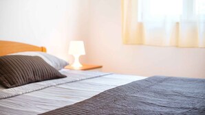 A neatly made bed with a striped bedspread and pillows, a bedside lamp, and a window with curtains. - Holiday Apartment Marina 5 2+1 (Srima)