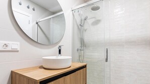 A modern bathroom with a round mirror, a white sink, wooden cabinetry, and a glass-enclosed shower. - GuestReady - New apartment in a quiet area (Valencia)