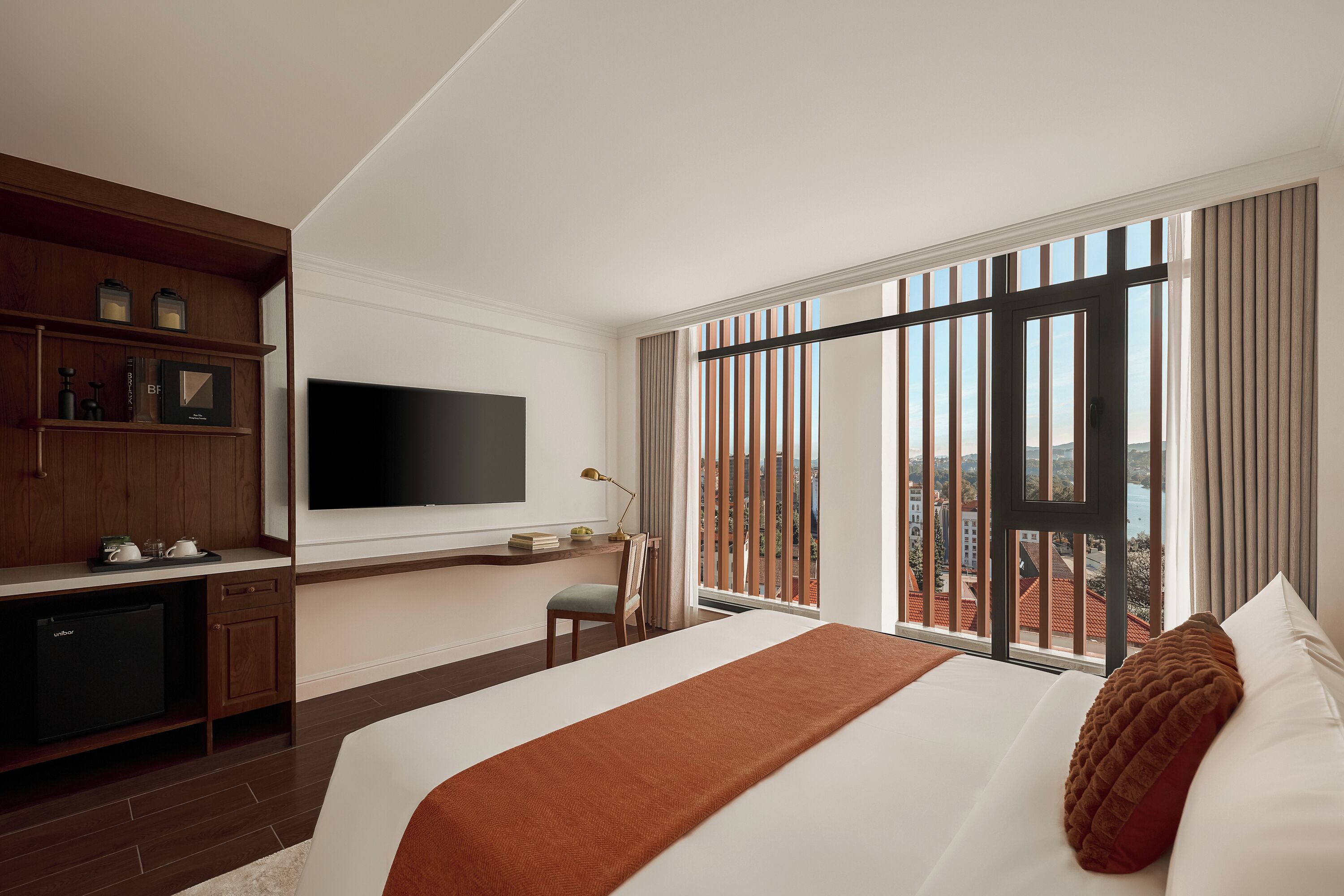 Habitación de hotel con cama, escritorio con televisor de pantalla plana, minibar y vistas a la ciudad a través de amplias ventanas.