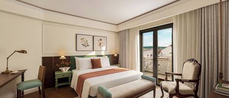 A hotel room with a large bed, a desk, a chair, a nightstand, a lamp, and a view of the outside.