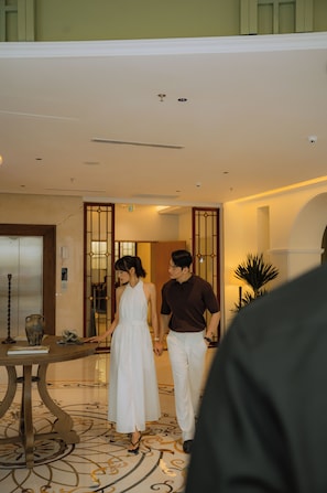 A couple walking hand in hand in a lobby with a patterned floor, a reception desk, and a door with a traditional design.