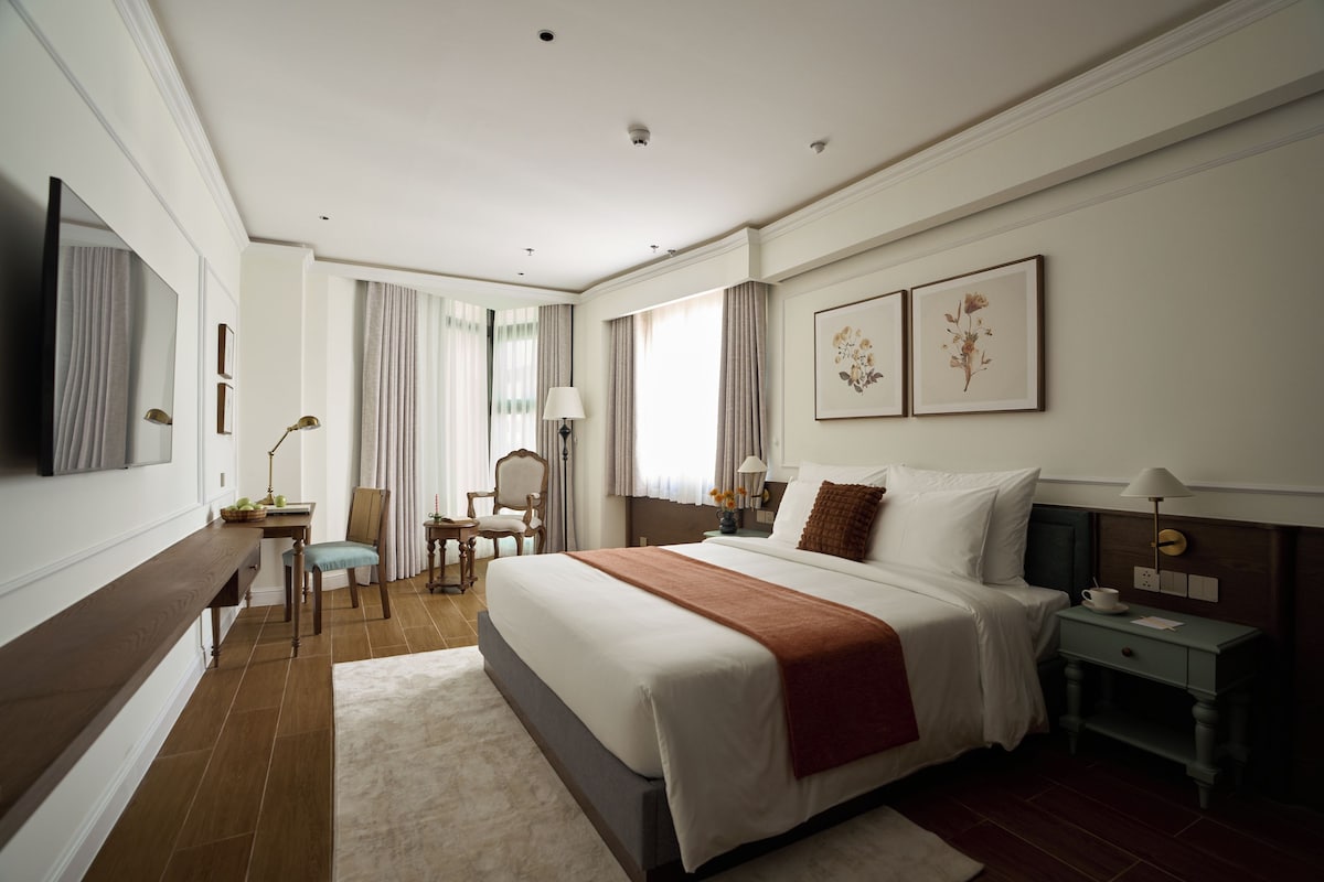 Quarto de hotel com uma cama grande, escrivaninha, cadeira, televisão e obras de arte nas paredes.