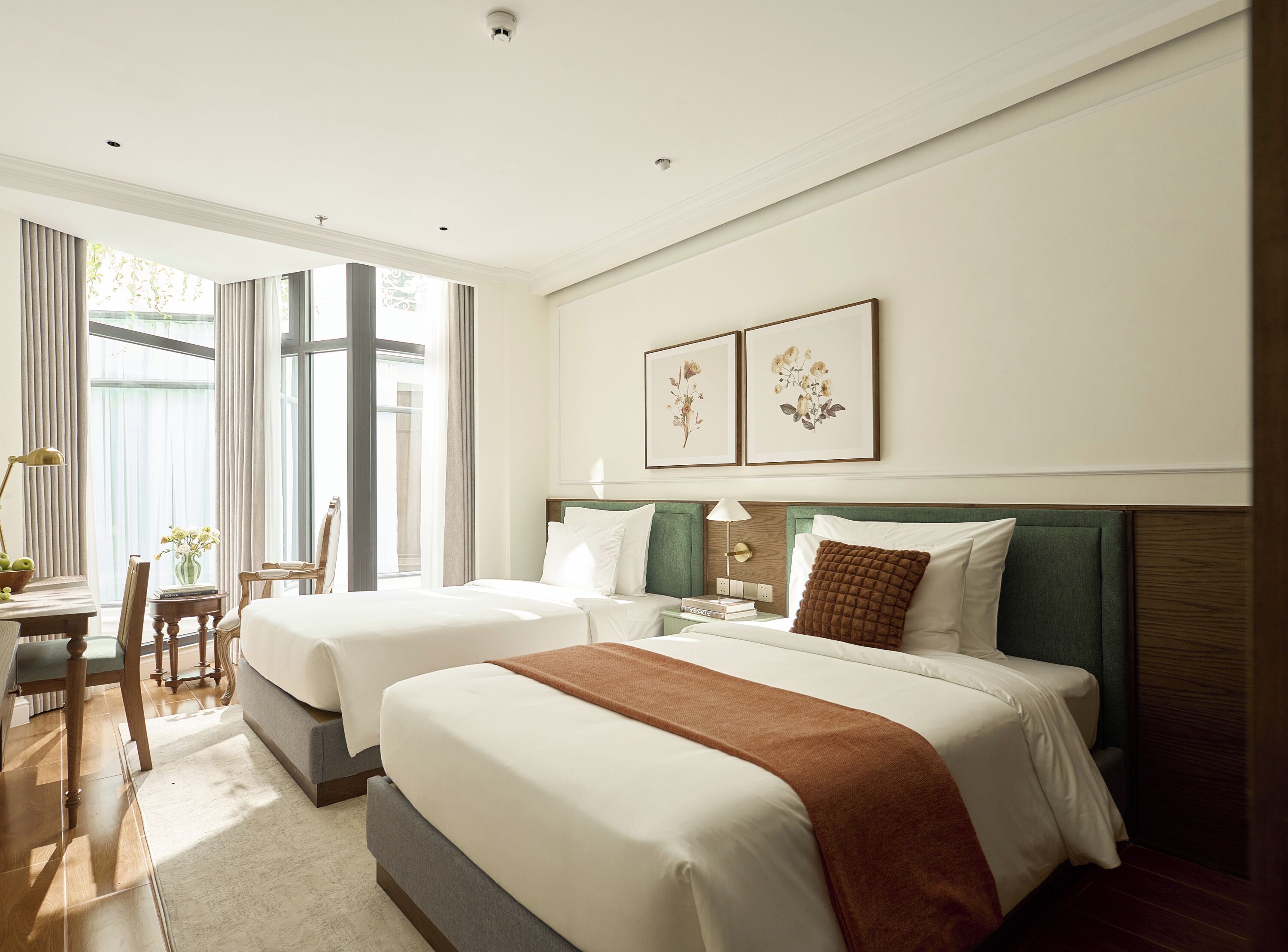 A hotel room with two beds, a desk, and a chair. There are two framed pictures on the wall.