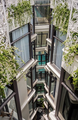 Un atrio de varios niveles con una escalera central, rodeado de balcones con barandillas de hierro blanco y plantas trepadoras verdes.