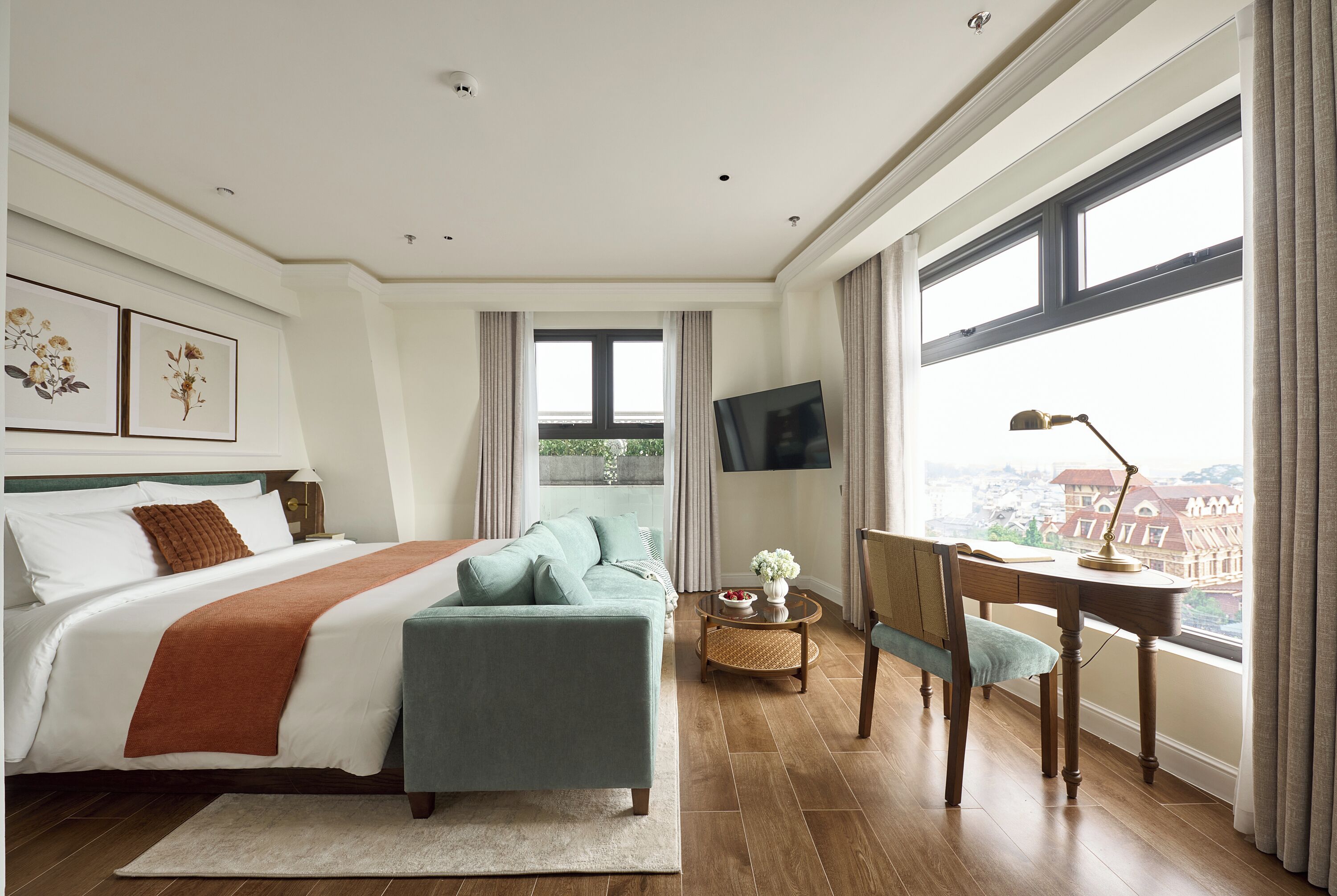A bedroom with a bed, a desk, a chair, a TV, and a window with a city view.