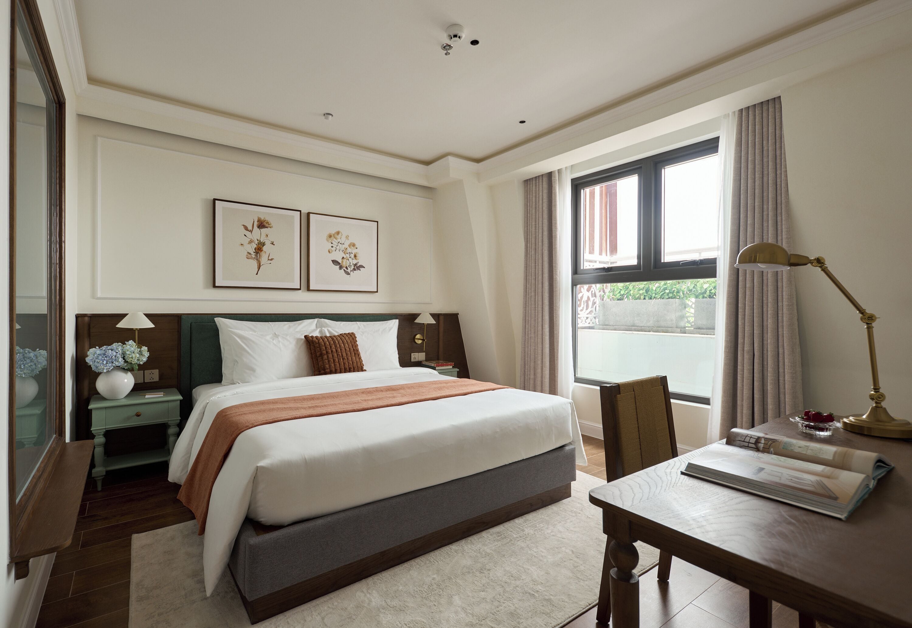 A hotel room with a large bed, a desk with a lamp, a window with curtains, and two framed pictures on the wall.