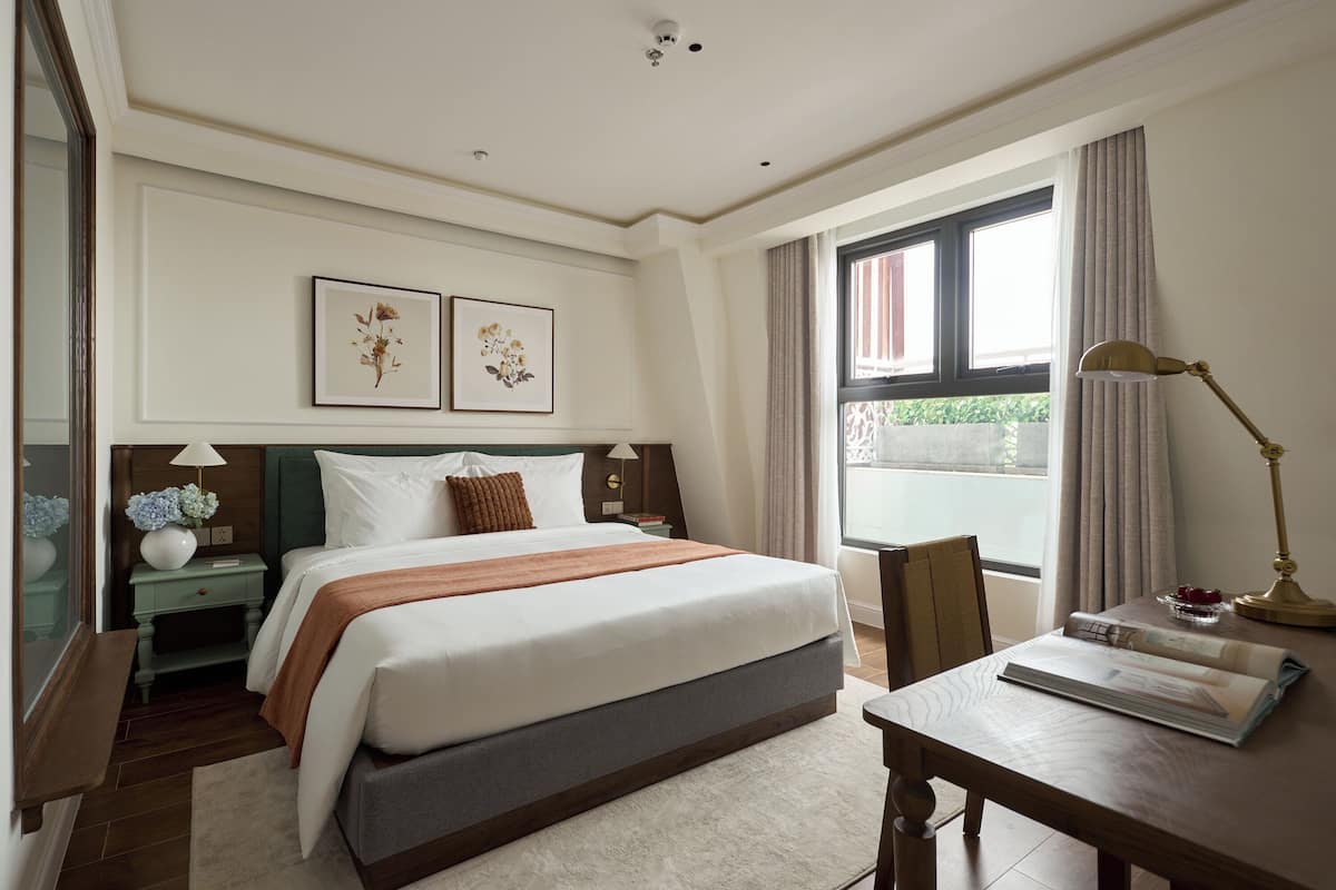 Une chambre d’hôtel avec un grand lit, un bureau avec une lampe, une fenêtre avec des rideaux et deux tableaux encadrés au mur.