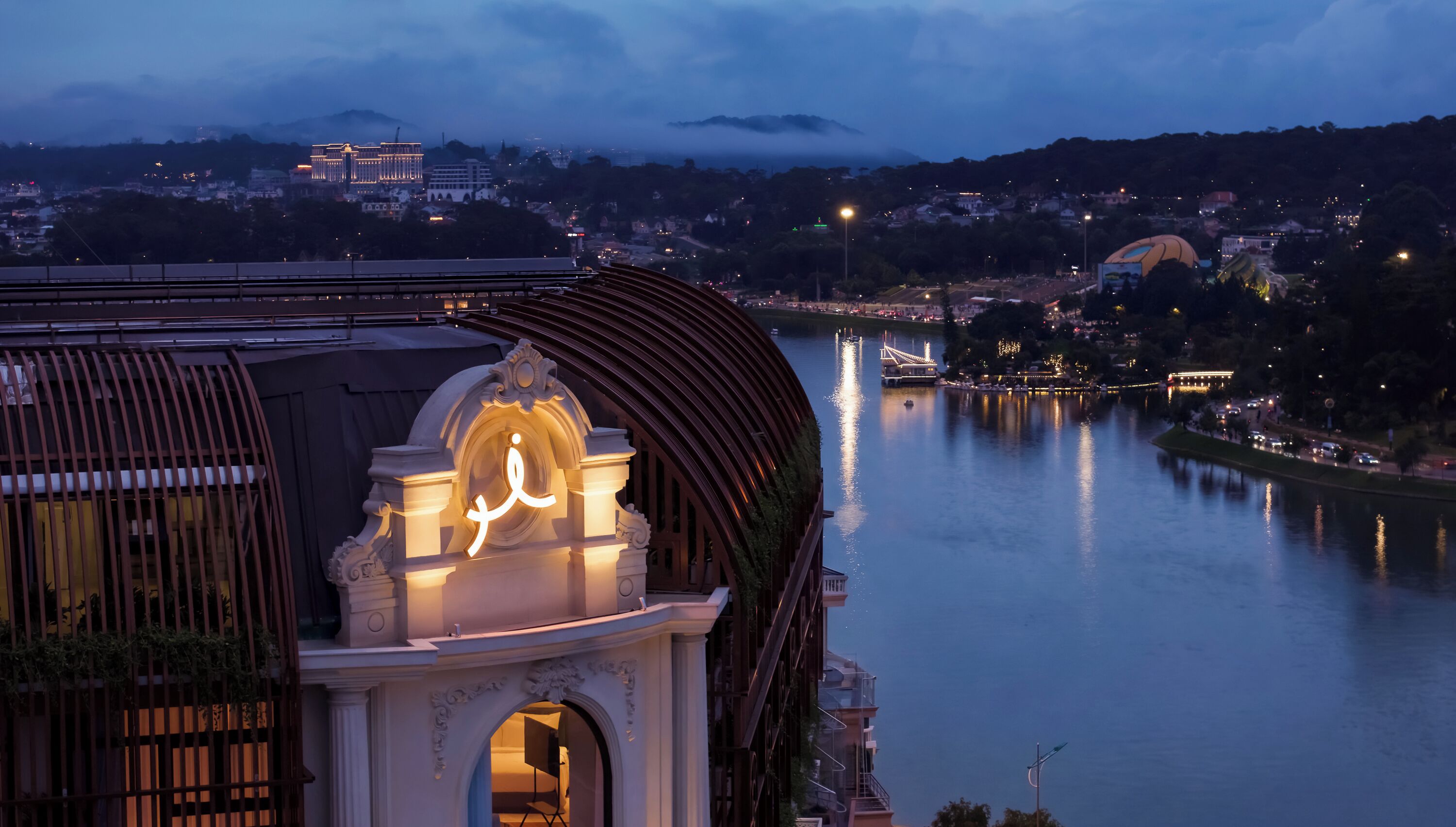 A building with a prominent archway and illuminated signage, overlooking a river with a bridge and distant cityscape.