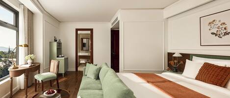 A modern hotel room with a green sofa, a bed with white linens, a view of the city, and a desk with a lamp and flowers.