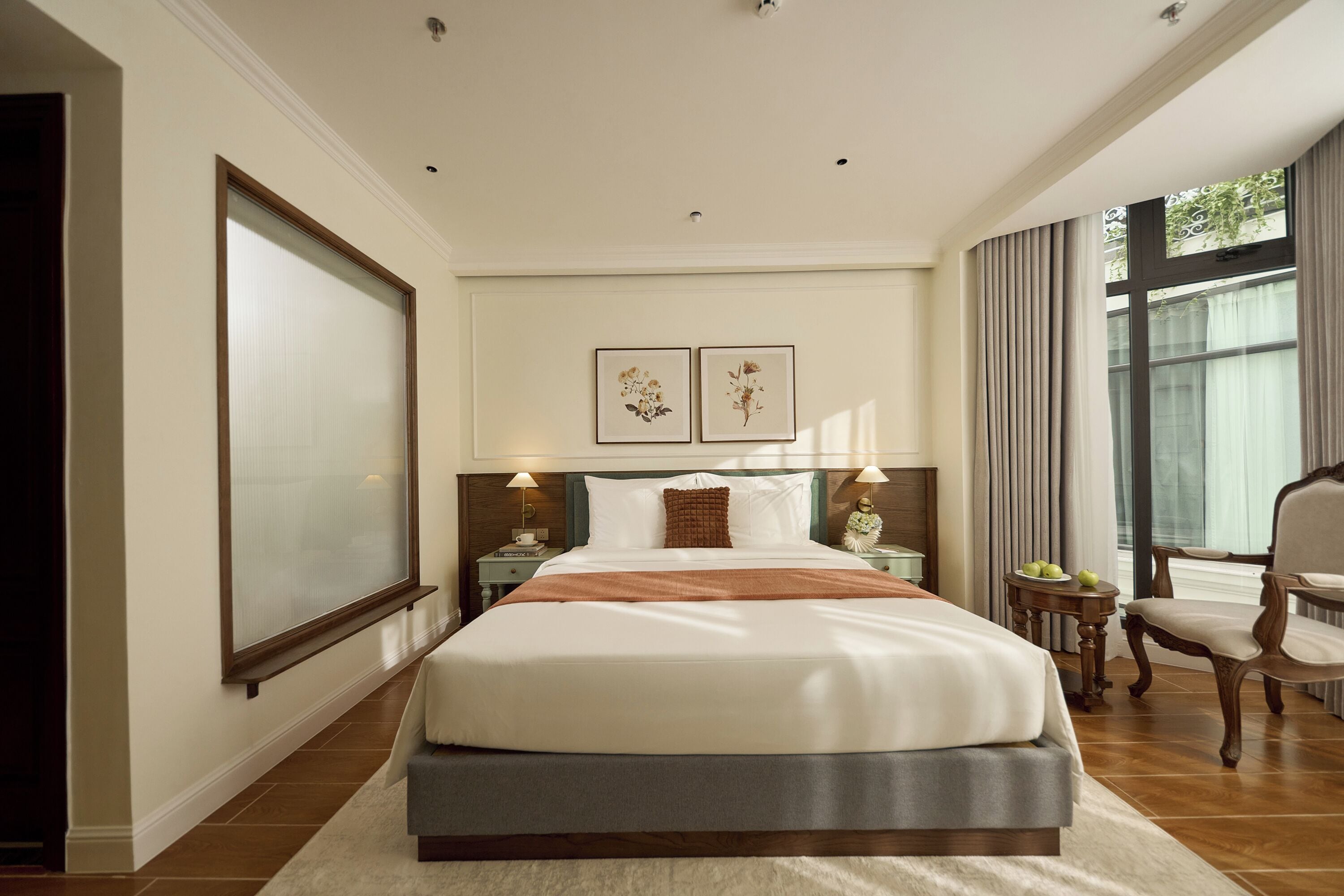 A hotel room with a large bed, two bedside lamps, a large window with curtains, and a view of greenery outside.