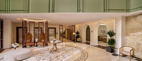 A spacious lobby with a high ceiling, a central seating area with chairs and a table, and a decorative floor pattern.