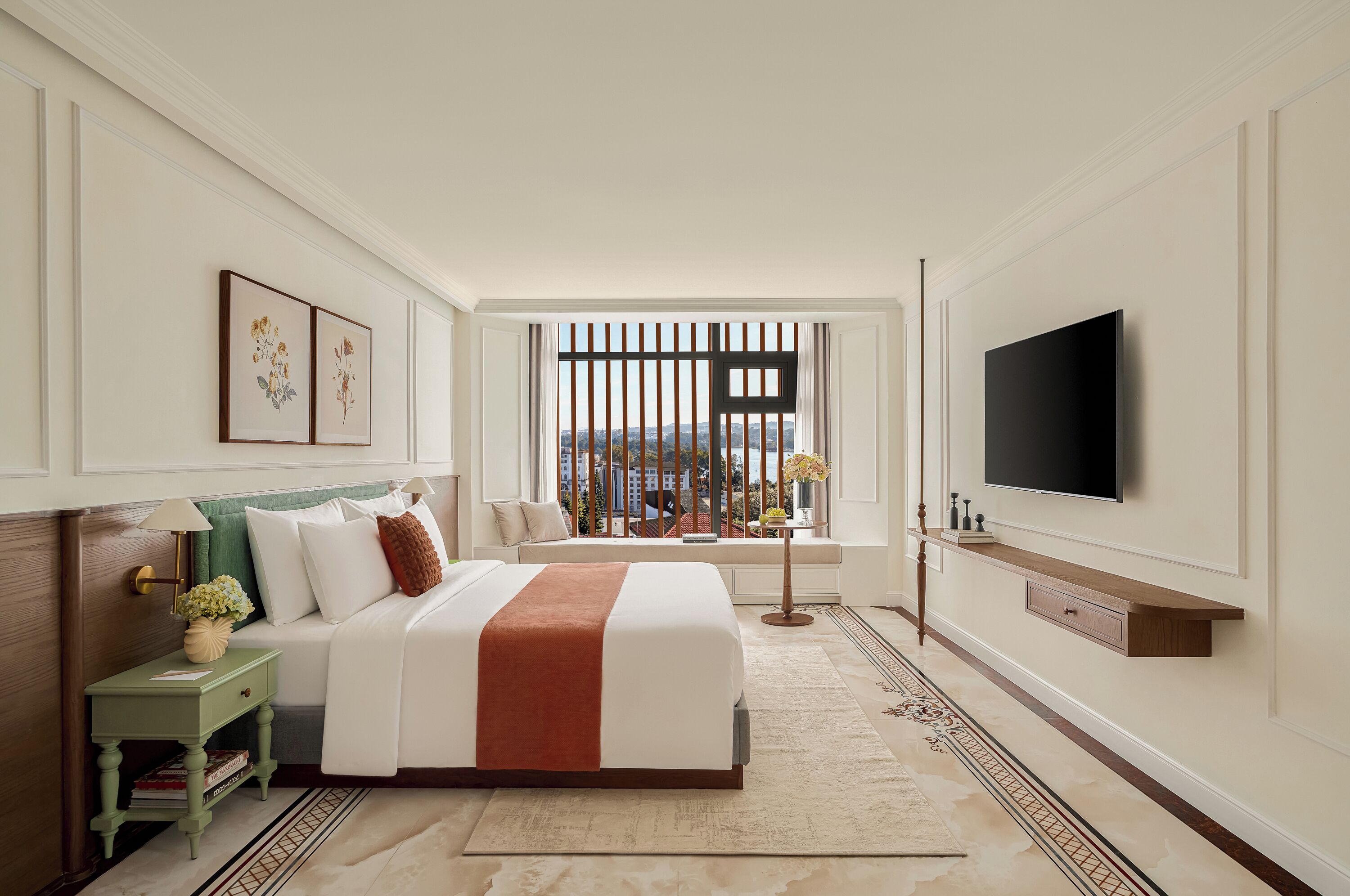 A hotel room with a large bed, a TV mounted on the wall, a window with a view of buildings, and a bedside table with a lamp.