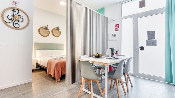 Una habitación de hotel moderna con cama, zona de comedor con mesa y sillas, un reloj de pared y una puerta con señal de salida de emergencia.