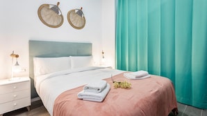 Un dormitorio con cama, dos mesitas de noche, cabecera, cortina color turquesa y dos cestas fijadas a la pared.