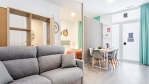 Una sala de estar moderna con un sofá gris, una zona de comedor con mesa y sillas, y una puerta con cortina verde.