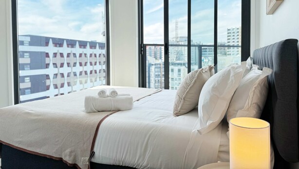 Ein Hotelzimmer mit einem großen Bett, weißen Leinentüchern, einer Nachttischlampe und Blick auf die Stadt durch große Fenster.