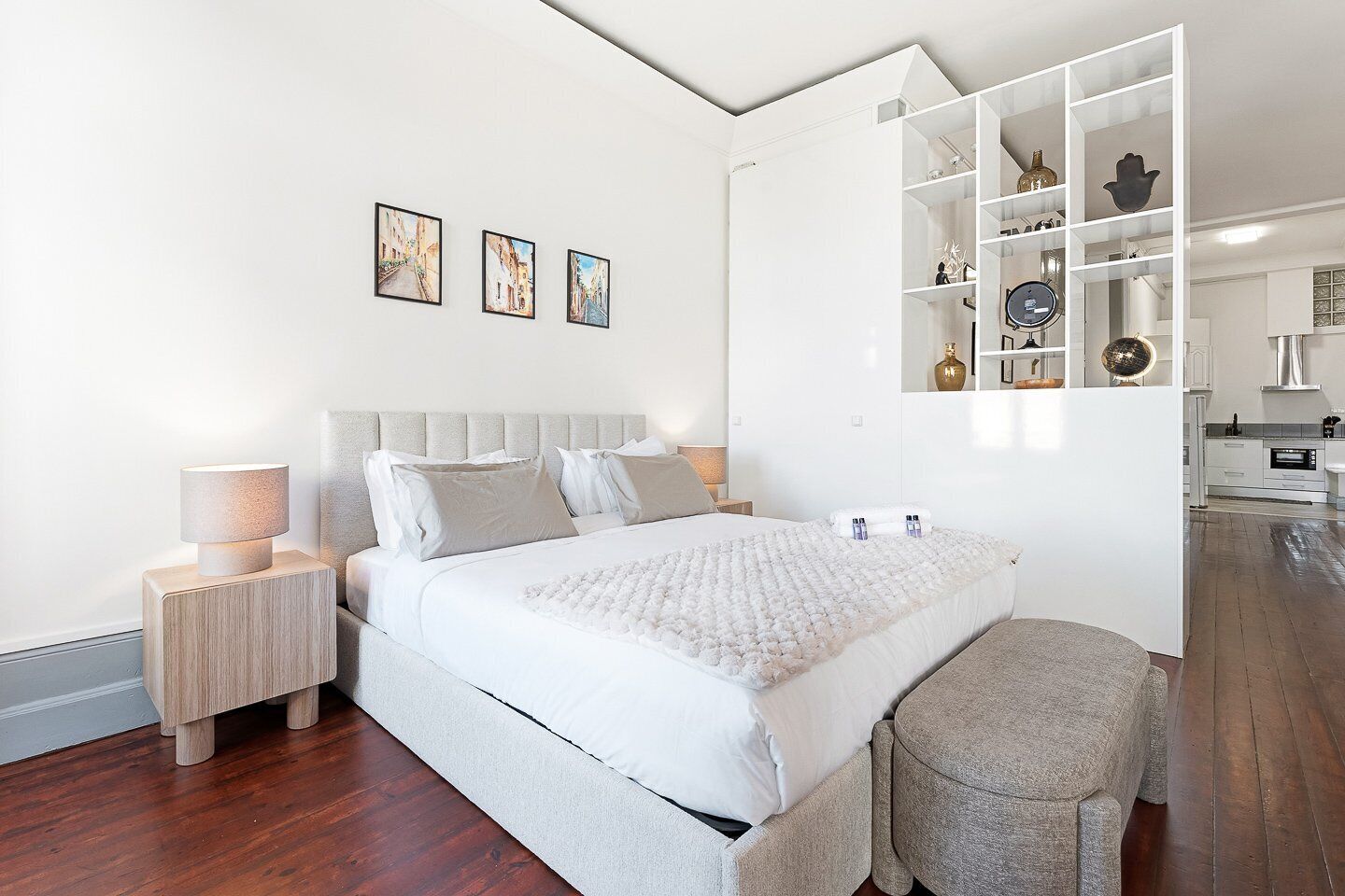 A modern bedroom with a bed, bedside tables, a bench, and a built-in white shelving unit with various items.