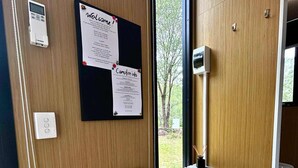 A room with a wooden wall, a bulletin board with welcome and campfire info, a bench, and a table with a view outside. - Mirror Reflections 2 by Tiny Away (Tamborine)