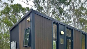 A small, modern cabin with a wooden exterior, a sliding door, and a small deck with steps leading to a picnic table. - Mirror Reflections 2 by Tiny Away (Tamborine)