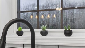 A kitchen with a black faucet, three potted plants on a white ledge, and a window with hanging lights. - Modern Retreat w/ Private Hot Tub & Bunk Room (Jasonville)