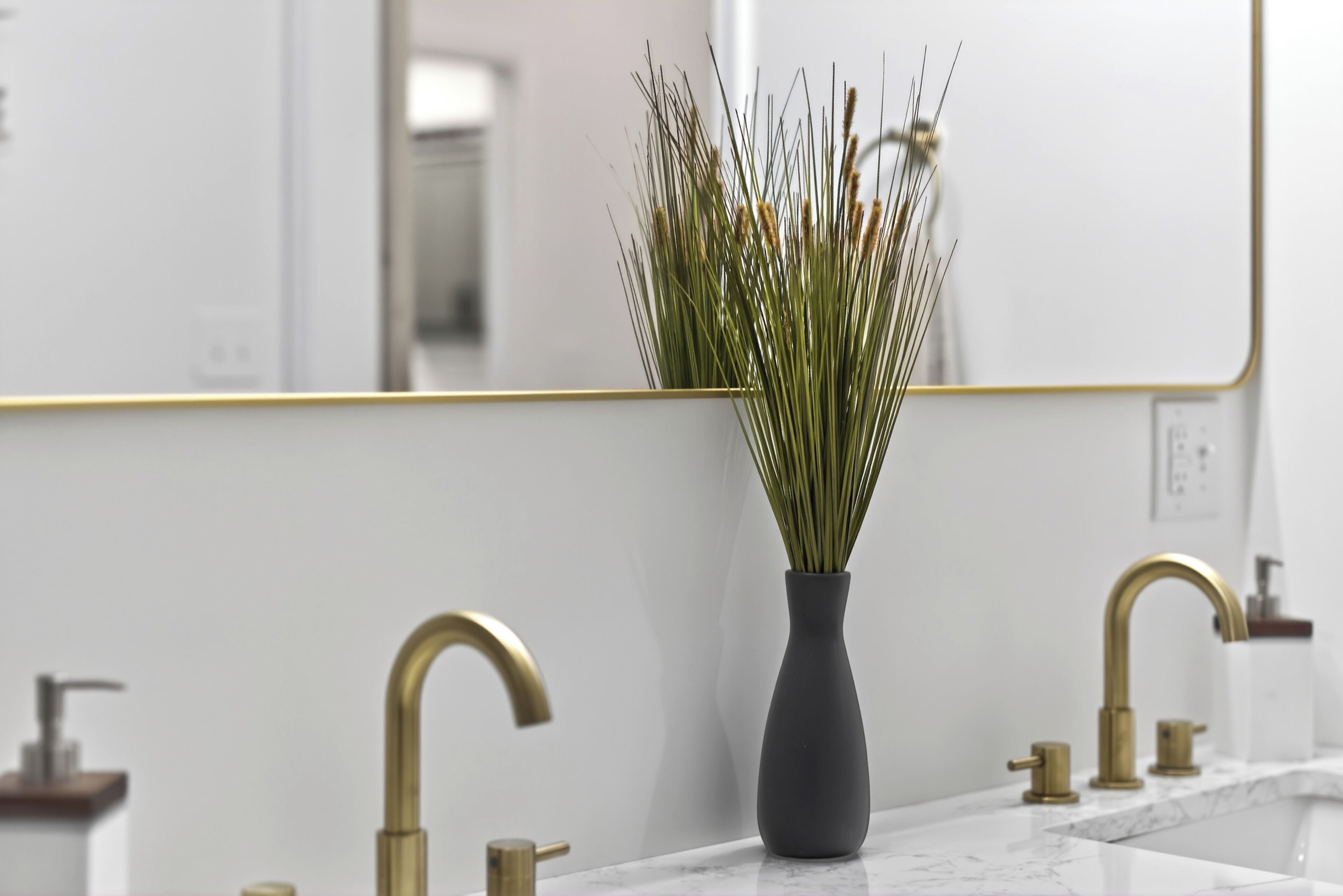 A bathroom with a white marble countertop, a dark vase with green plants, and gold faucets.