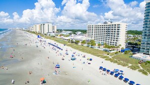 Una playa con muchos sombrillas y gente, un hotel grande con varios pisos y un cielo despejado.
