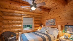 A log cabin bedroom with a bed, a chair, a nightstand, a ceiling fan, a window, and a painting on the wall. - Otter Point Lodge - 4BR, 3BA 1100ft water frontage log home w/ 2 docks & firepit (Land O' Lakes)