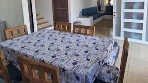 A dining area with a table covered in a patterned tablecloth, wooden chairs, a small white table, and a blue sofa in the background.