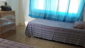 A room with two beds, a wooden dresser, and a window with blue curtains.
