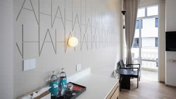 A modern hotel room with a minimalist design, featuring a geometric wall art, a dining area with a table and chairs, and a large window offering a view.