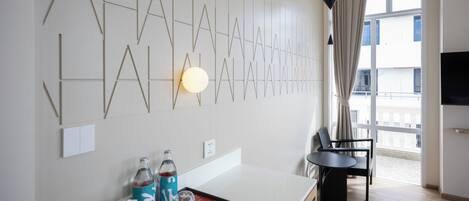 A modern hotel room with a minimalist design, featuring a geometric wall art, a dining area with a table and chairs, and a large window offering a view.