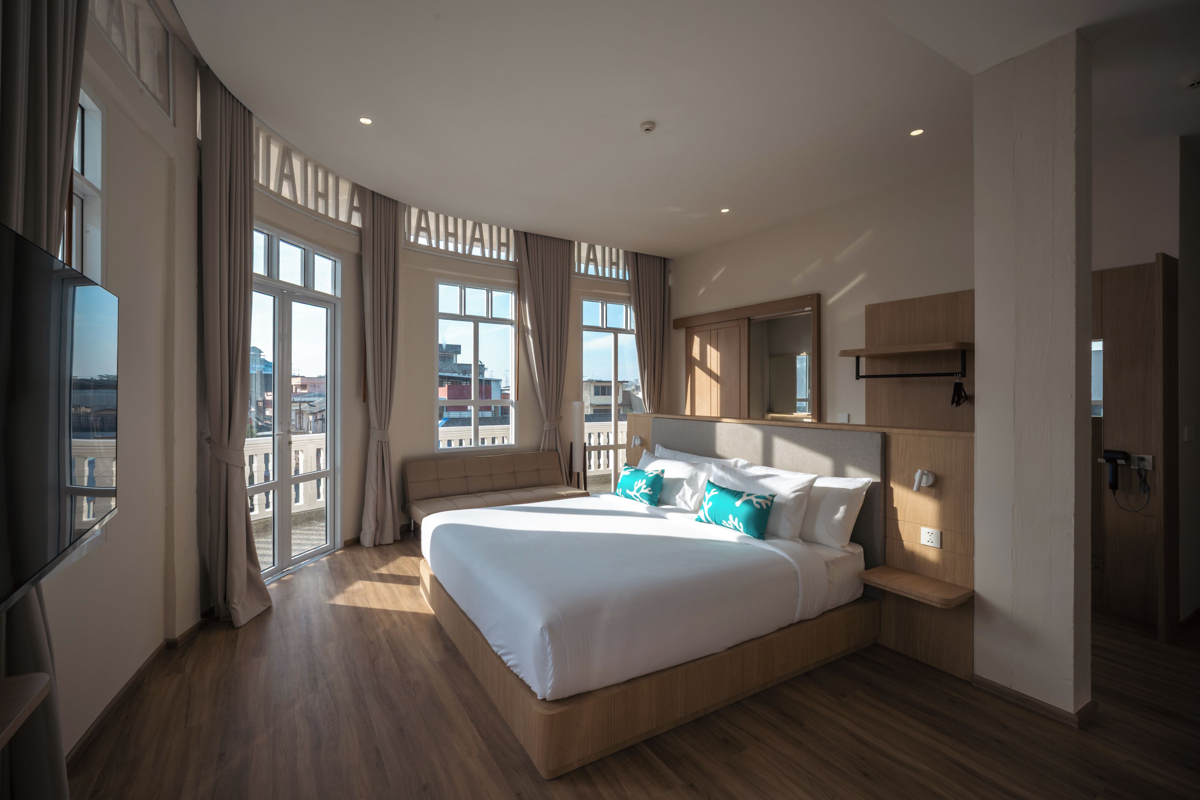 A modern hotel room with a large bed, a flat-screen TV, a wooden headboard, and a balcony with curtains.