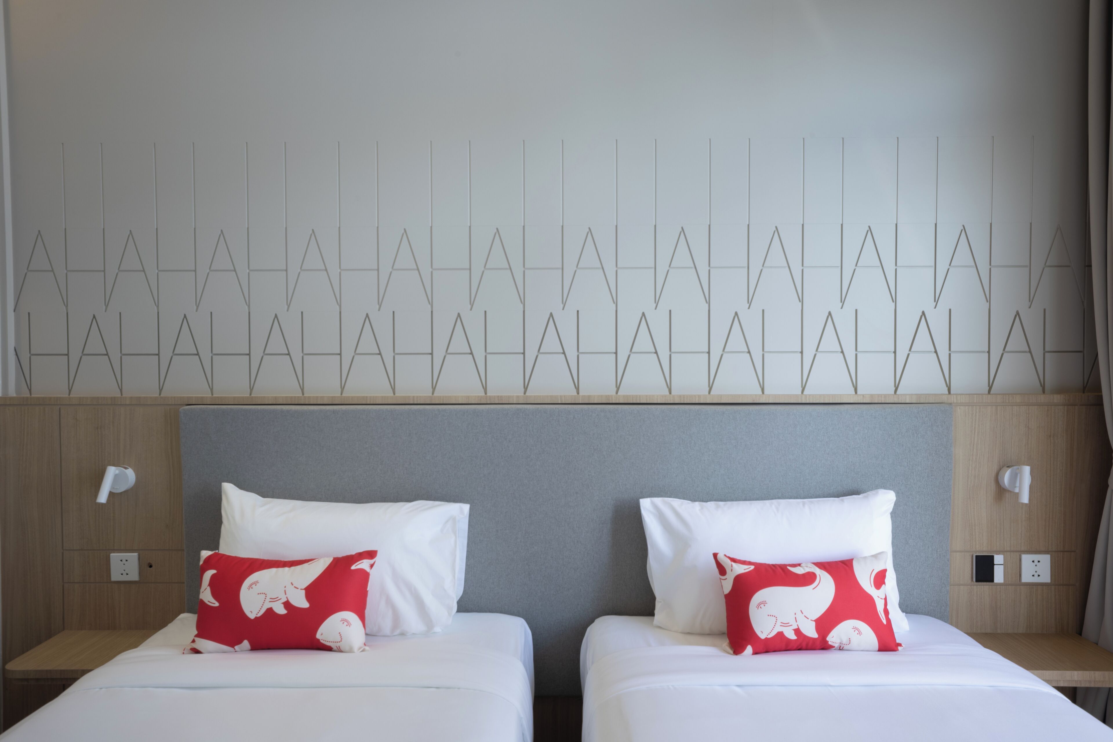 A hotel room with two beds, each with a white mattress and a red pillow with a white elephant design. The headboard is grey, and the wall features a geometric pattern.