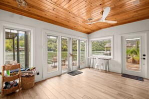 Una habitación amplia con piso de madera, ventanas grandes, ventilador de techo y vista a un lago.