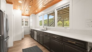 A modern kitchen with wooden ceiling beams, white cabinetry, a stainless steel refrigerator, and large windows offering a view of a staircase and greenery. - Lake Austin Bungalow | Private Dock + Swim Off the Dock (Austin)