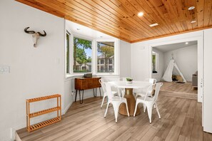 Un interior moderno con una mesa redonda de comedor, sillas blancas, techo de madera y ventanas amplias.
