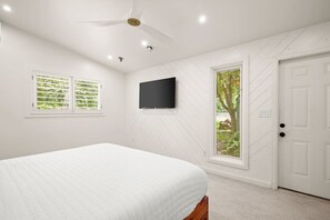 Un dormitorio con cama, ventilador de techo, televisor y dos ventanas con persianas.