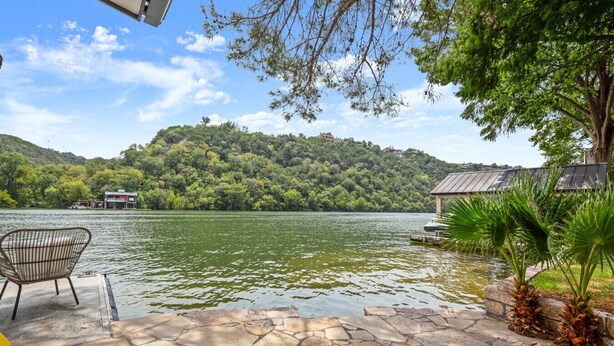 A stone deck with a chair, a small boat, and a gazebo by a lake with a forested hill in the background. - Lake Austin Bungalow | Private Dock + Swim Off the Dock (Austin)