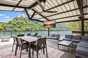 A covered outdoor area with a table and chairs, a sofa, and a TV screen displaying a sunset view.