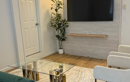 A modern living room with a green sofa, a glass coffee table, a white armchair, and a large flat-screen TV mounted on a light-colored wall.